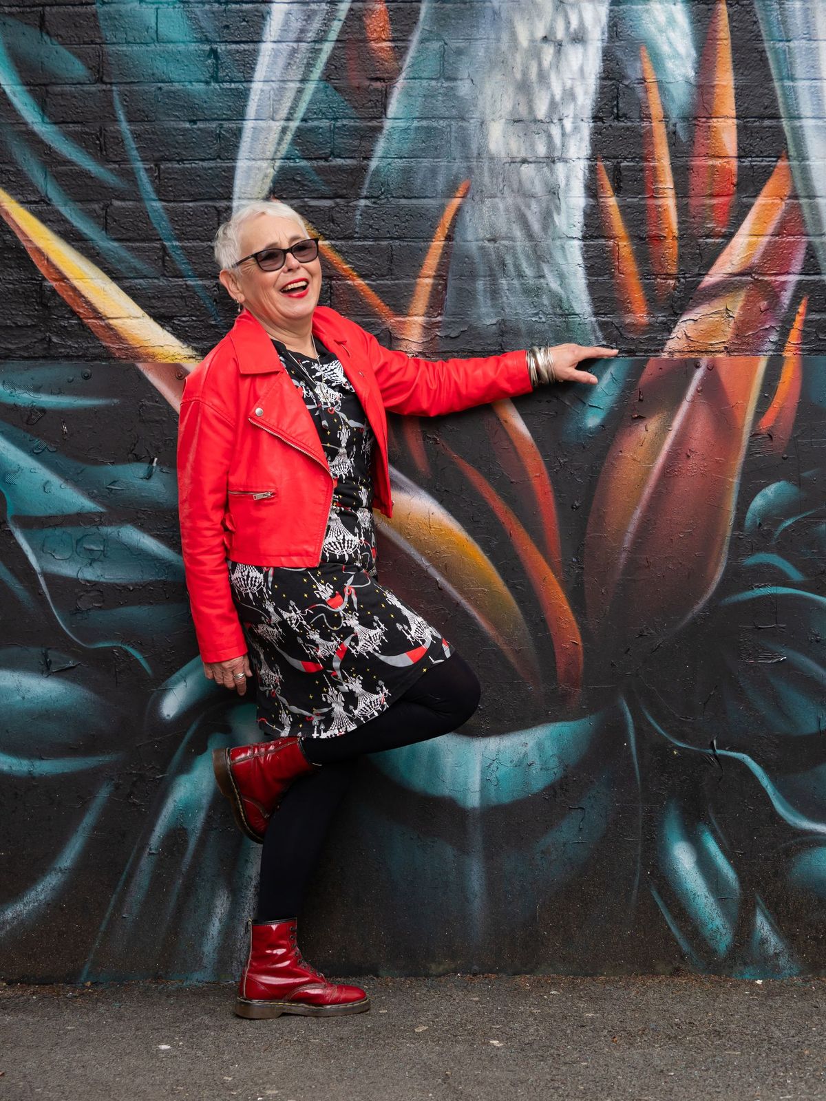 A smiling older woman in a bright red jacket in front of colourful street art