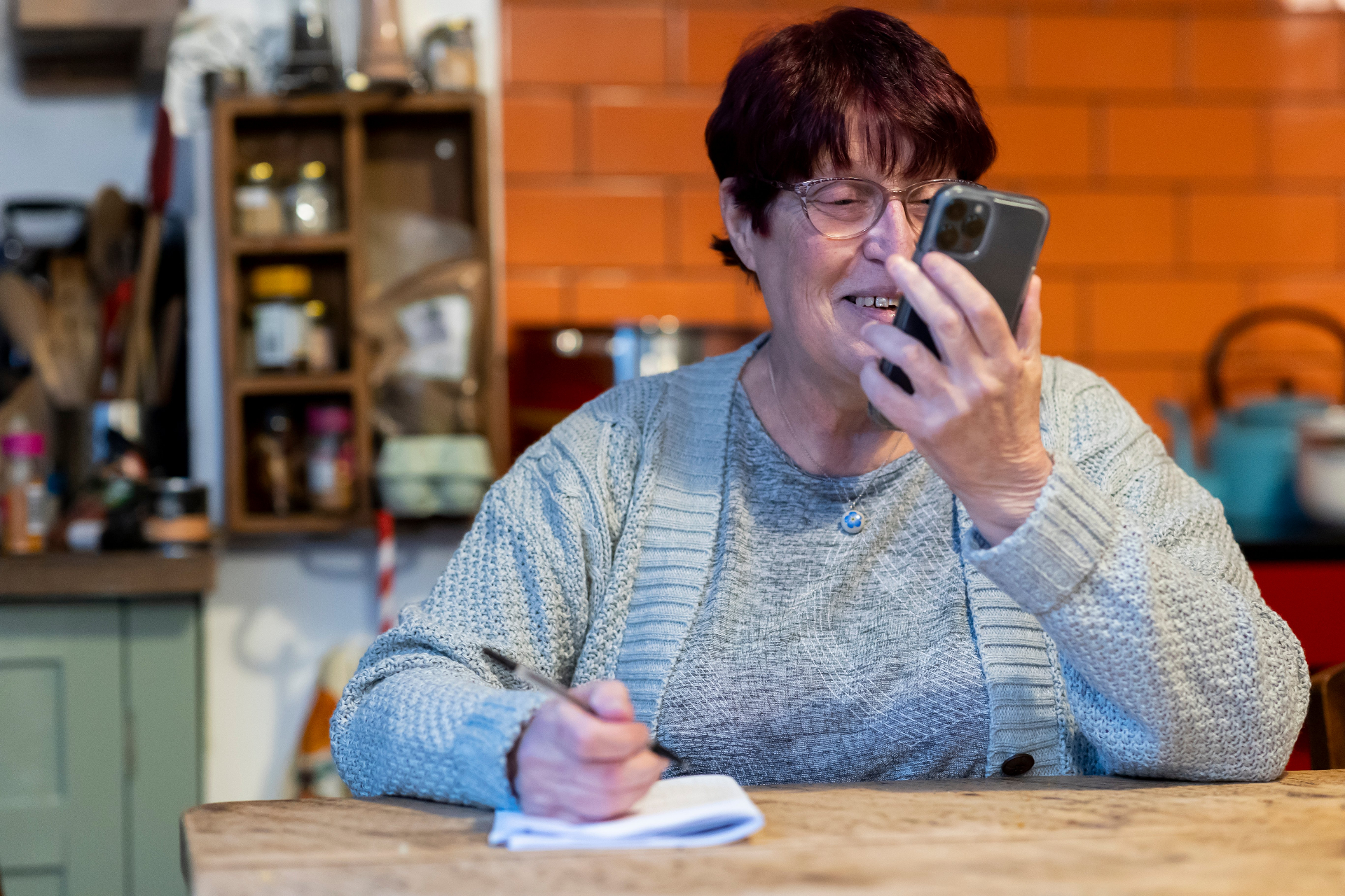 An older woman smiling while using her phone and taking notes at her kitchen table