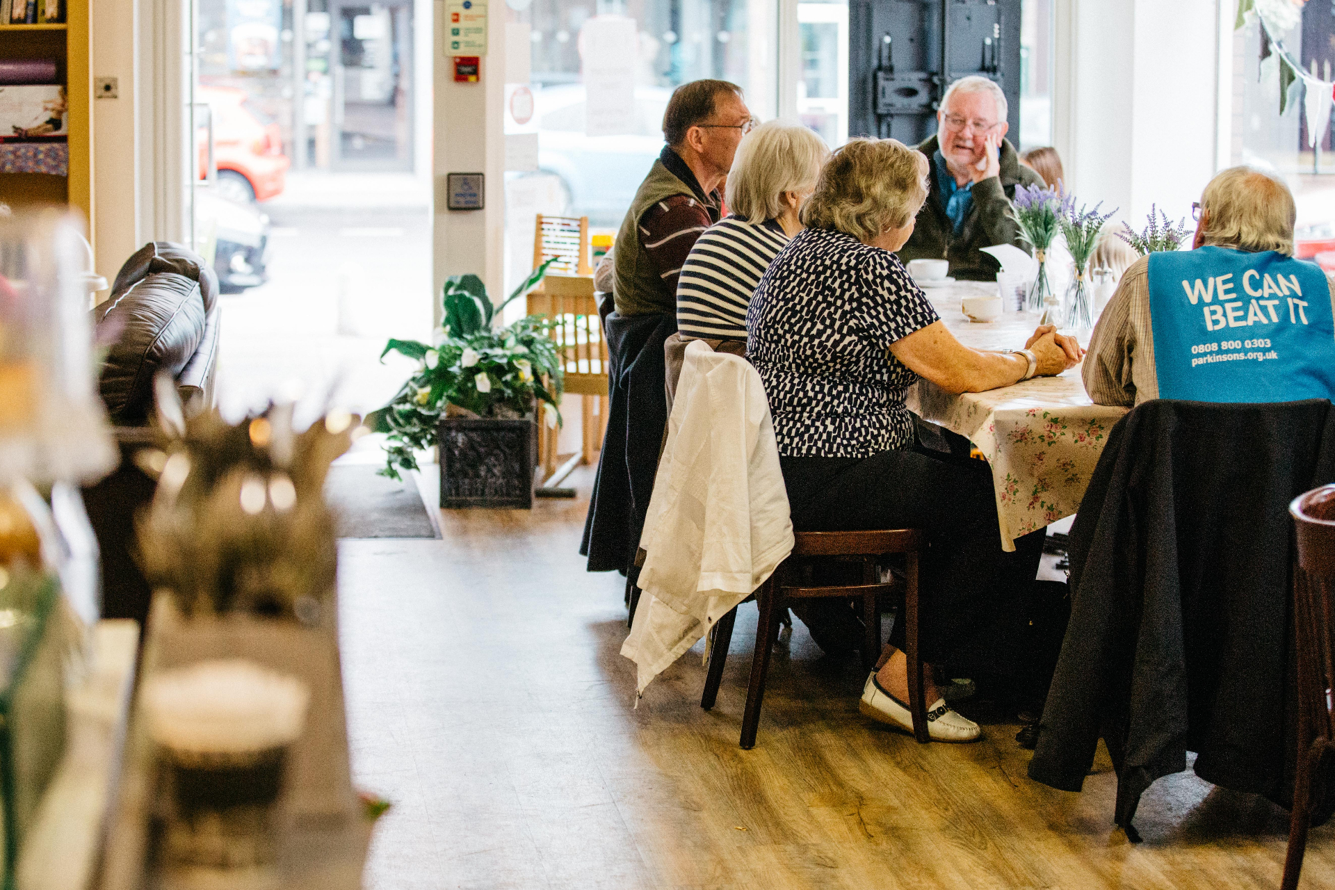 Older guests chatting around a table at Communitea Café with a Parkinson's UK 'We Can Beat It' tabard visible