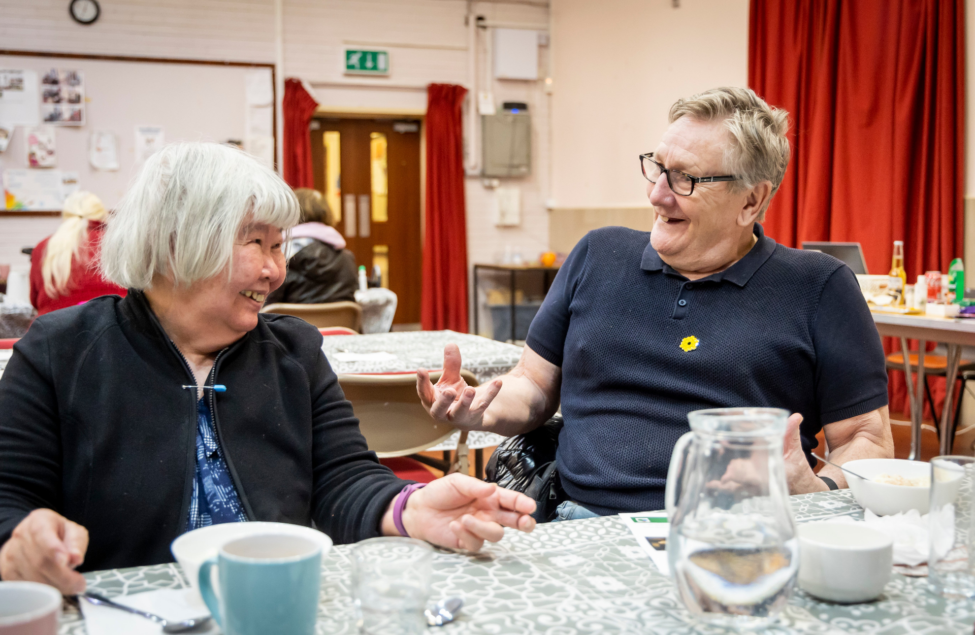 Two older people chatting and laughing together at a community centre table.
